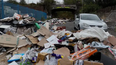 Heaps of rubbish including cardboard boxes, debris and cloth have been dumped along a road. A car is parked nearby and there are blue railings. There is a bridge at the end of the road, which is closed off with gates.