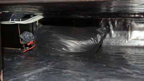 West Yorkshire Police The interior of a concealed storage compartment or cavity, viewed through a low opening.
In the centre of the image is a large black plastic-wrapped package. The package appears tightly wrapped and elongated, with a rounded shape. On the left end of the package, a section of the wrapping is gathered and secured, and a small red strap or tie is visible.