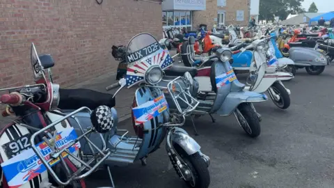 A row of several Lambretta scooters, without their riders, are lined up in a car park