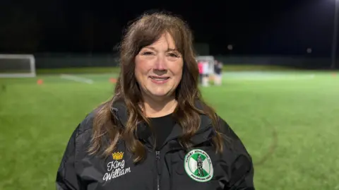 Denise stands smiling at a training session wearing her black Tunley Athletic-branded coat