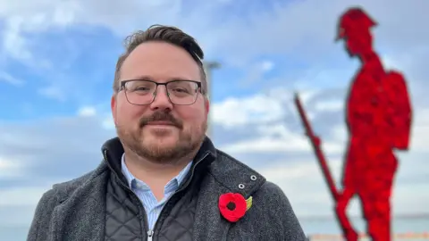 Daniel Cowan is wearing a grey jacket over a black coat and pale blue shirt. He has a crocheted poppy pinned to his jacket. He is smiling. In the background is a red statue of a soldier.