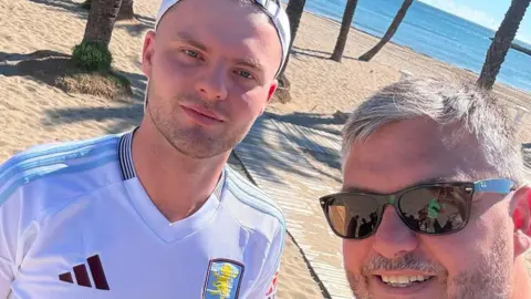Two men standing on a beach with trees and the sea behind them. One is wearing an Aston Villa top with white backwards cap looking at the camera. The one is wearing sunglasses with grey short hair and a beard. 