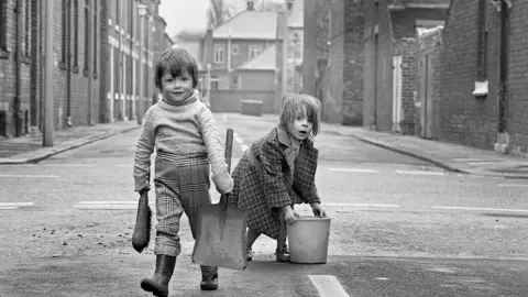 A black and white image shows two children in an empty terraced street. One is carrying a shovel and brush the other is bent over lifting a bucket. The first has check trousers tucked into wellies and a thick jumper which looks a little too big. The other wears a long, warm check coat. Both are looking into the camera.