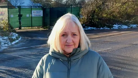 Sue Bussey stands in a blue coat with the entrance to the proposed site behind her