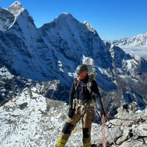 Mid and West Wales Fire Rhys Fitzgerald standing on a mountain, while holding a rope. In the background are higher mountains, and the sky is a cloudless blue