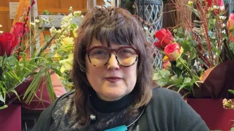 Kathryn Hulme mid-length brown hair, wearing large tortoiseshell glasses, looking at the camera and surrounded by flowers in the florist business that she owns