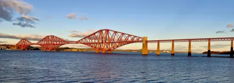 Erin Harper The Forth Rail Bridge over the Firth of Forth. The bridge has three cantilevers and is painted red. The water has ripples in it and there are a few clouds in the sky.