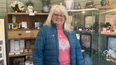 A woman, with shoulder length blonde hair wearing a pink jumper and blue coat and glasses. She is wearing a crystal pendant necklace. Behind here are crystals inside glass shelves.