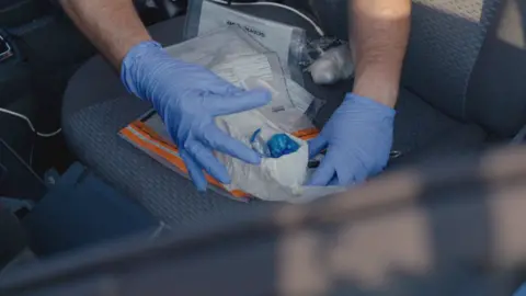 Bedfordshire Police Drugs in various plastic bags and wrappers are being examined by a man wearing blue gloves. It looks like they are on a seat inside a car