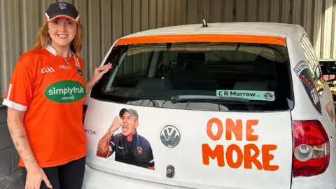 Gillian Kelly Gillian Kelly wearing an Armagh GAA top standing beside a white car that has been decorated with orange paint and a picture of the Armagh manager.