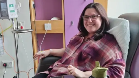 PA Media A woman with shoulder-length brown hair and glasses is smiling and sitting in a chair. She has a cannula in her hand which is wired into a machine. She is wearing a pink, purple and white plaid top.