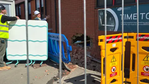 A damaged brick wall outside a building, cordoned off with safety barriers, with a hole in the wall and construction workers nearby.
