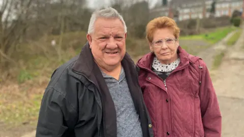 BBC/Simon Thake An elderly man in a black coat stands next to an elderly woman in a red coat with a stretch of waste land behind them.