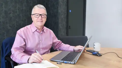 Middlesbrough Council A photo of Clive Heaphy, a grey haired man with glasses and a pink shirt sitting in front of a laptop, smiling at the camera.
