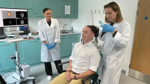 A demonstration with a colleague showing how gel is first placed on the head before an ultrasound transducer is held against the scalp. There are two researchers, with another member of staff demonstrating the process for illustrative purposes.