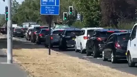SHAM ASHRAF Daytime traffic jam on a road, showing a long queue of cars and a white van stationary near traffic lights, with blue road signs for bus and cycle lanes visible in the foreground.