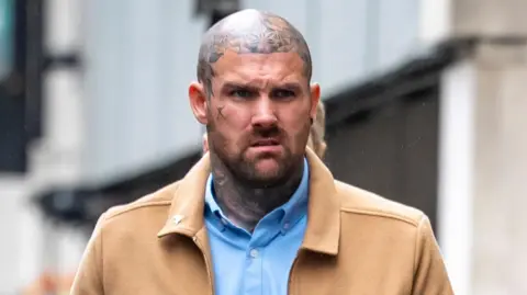 Tony Bartlett walks towards Bristol Crown Court. He is a large man with a shaved head and several tattoos on his neck and head, but not on his face itself. He is wearing a light blue shirt under a light brown jacket.
