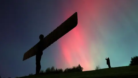 The Northern Lights glowing red and lighting up the Angel of the North, as someone standing behind takes a picture.