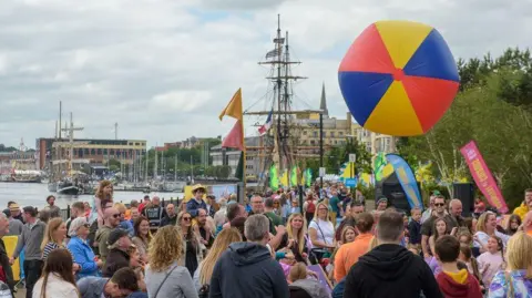 DCSDC a large crowd of smiling people gather on the quay in Derry enjoying the martime festival's activities. A large, inflatable ball which is yellow, blue and red is floating in the air while ships and the River Foyle can be seen in the background