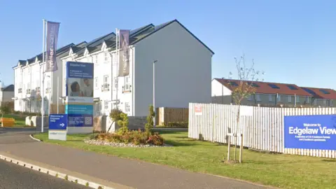 Entrance to Miller Homes' Edgelaw View development, showing blocks of flats, landscaped grounds, roadside signage and a residential street under a clear blue sky.