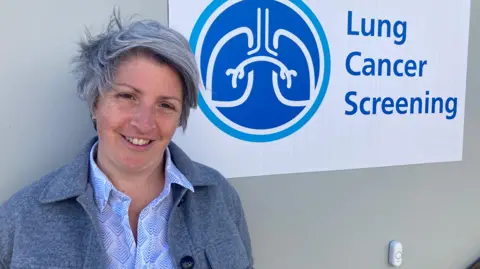Dr Anna Bibby wearing a light shirt and grey jacket standing next to the unit which has a lung cancer screening sign on the side.