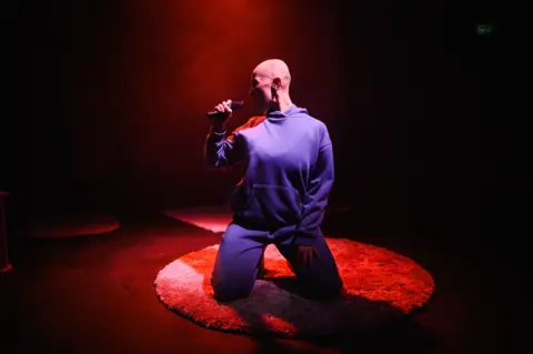 Charlie Flint Emily Glaze kneels on stage and holds up a microphone to her mouth. She wears a blue tracksuit and is kneeling on a rug.