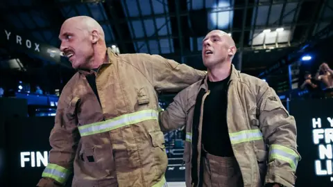 Kieran Thomas Kieran and Andy embrace as they cross the finish line at the Hyrox in Manchester. They're red faced and still wearing the firefighting kit.