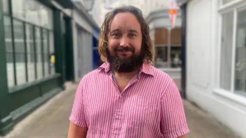 Man with sholder length brown hair and pink and white stripy shirt.