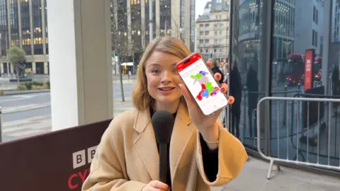 Megan Davies in a cream coat with blonde hair holding a phone which shows the constituencies