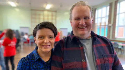 Leah Calvert has short brown hair and is wearing a blue and black blouse. She stands beside her husband Geoffrey who is taller and has grey hair. He is wearing a blue and red checked overshirt over a grey T-shirt.