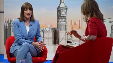 Rachel Reeves sits on a red armchair and wears a blue suit and sits opposite Laura Kuenssberg who is seated on a red armchair in a red dress, while appearing on the BBC 1 current affairs programme, Sunday with Laura Kuenssberg.