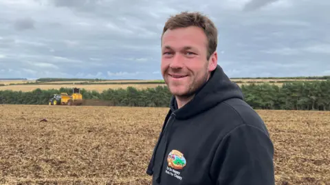 Ben has short brown hair, a beard and is wearing a black hoodie. He is standing in a farmer's field and is smiling at the camera. 
