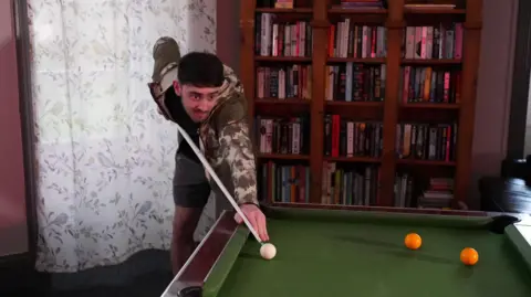 A young man with dark brown hair and a short brown beard and moustache is about to take a shot at a pool table. He is holding a cue with his right arm pulled back and the end of it is just touching the white ball. Two yellow balls are on the table to the right. The man is wearing grey shorts and a black t-shirt with an olive green camouflage patterned tracksuit jacket open over it. Shelves filled with books are visible to the right of the picture, and a long window covered with a white curtain patterned with birds and leaves is to the left. 