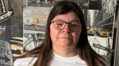 Hayley Ellis A woman with long brown hair, dark framed glasses and a white tshirt in front of a wall with black and white photos of streets on it