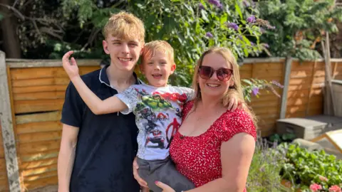 Becki Bowden / BBC A woman stands alongside her eldest son, whilst picking up her youngest son.