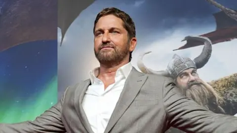 Gerard Butler wearing a grey suit jacket and a white shirt. He has dark hair and facial hair. Behind him is a poster for the film.