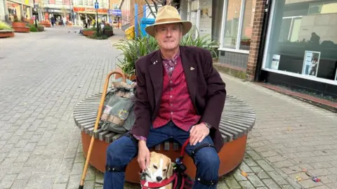 Kate Bradbrook/ BBC A man in a Purple blazer, red waistcoat, jeans and a beige hat sits on a circular bench in a town centre while petting his brown and white dog. A curved walking stock and a grey bag are next to them.  