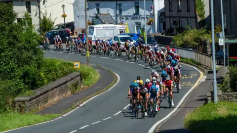 Getty Images A peloton of cyclists cycling round a corner on a Welsh road over a small stone bridge