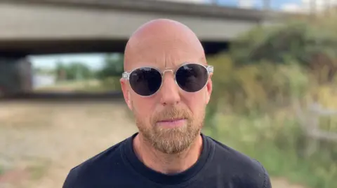 Luke Deal/BBC A man with a bald head, light brown beard and dark glasses and wearing a black t-shirt