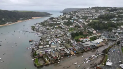 This is an aerial image of Salcombe, a seaside town. Boats are in the water. Colourful houses are visible. Sandy beaches can be seen on the left.