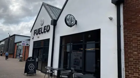 The outside of Fueled coffee shop. it is a primarily white building with Fueled in big letter across the top of the front door. There is also some seating out the front of the shop as well as an A-board used for advertising.