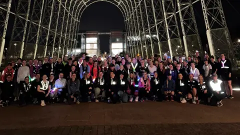 Jack Easton A large group of women pose together at night. They are wearing running gear and many of them have reflective tops. They are underneath a large metal arch and there is an illuminated building behind. 