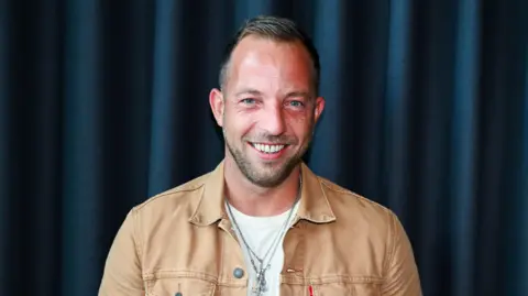 Musician James Morrison wearing a tan shirt and white t shirt. He has short dark hair and a short bead. 