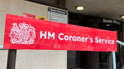 Lewis Adams/BBC A sign that reads HM Coroner's Service. It is a red sign and outside the coroner's court building in Chelmsford. 