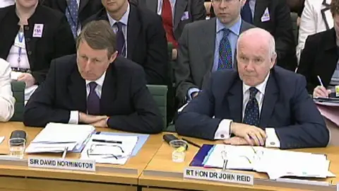 House of Commons Sir David Normington and John Reid seated at a table in a select committee, with papers spread out in front them, wearing pensive expressions