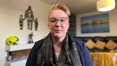 Shelley Martin - a woman with short blond hair and glasses - looks into the camera. She is wearing a blue jacket with a grey and black scarf wrapped around her.