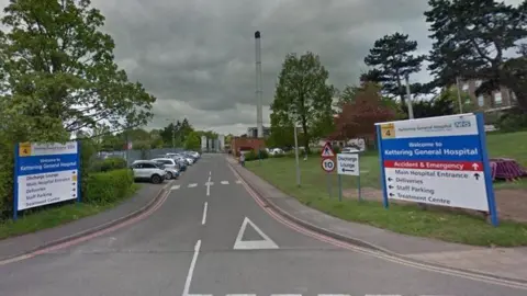 Entrance to Kettering General Hospital showing two-lane road heading past signs on either side, with black writing on a white background and a blue band towards the top on which Kettering General Hospital is written out in white lettering. There are cars parked in spaces on the left hand side of the road and trees to the left and right.