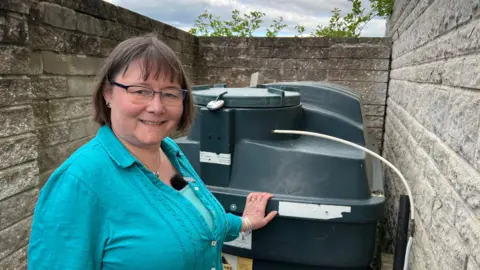 A woman with short dark brown hair and glasses stands next to a large green plastic tank.