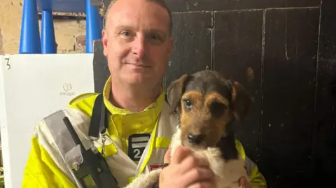 HIWFRS Firefighter holding a brown and white Jack Russell dog in his arms - the firefighters has short hair and is looking at the camera.