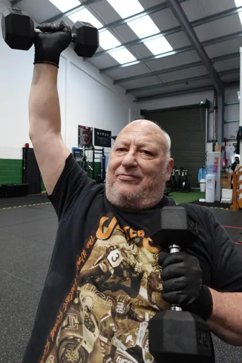 Patrycja Boryka/BBC A bald man with short grey stubble is smiling into the camera as one arm is lifted and the other is holding a black weight. He is wearing a black top with a picture on it and behind him is the empty floor of a gym.
